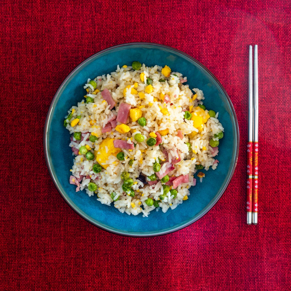 Overhead view of a blue bowl of fried rice with ham, peas, and corn, next to chopsticks.