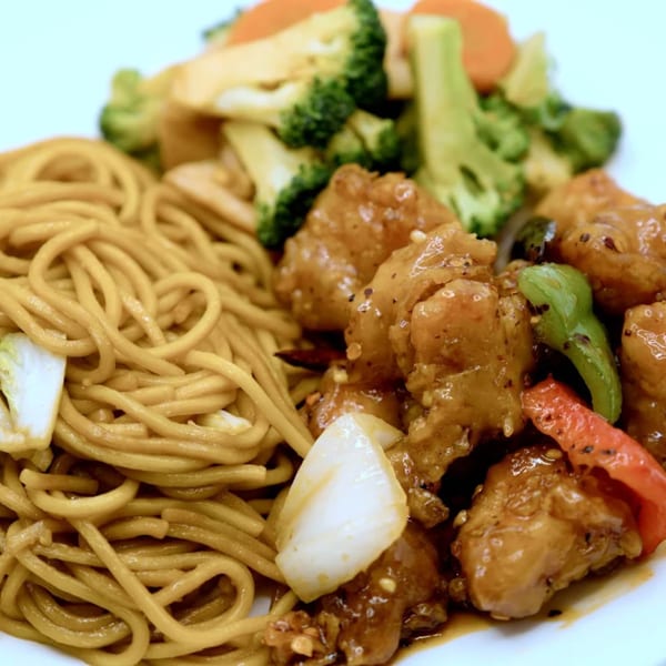 Sweet and sour chicken with lo mein noodles and steamed broccoli and carrots on a white plate.