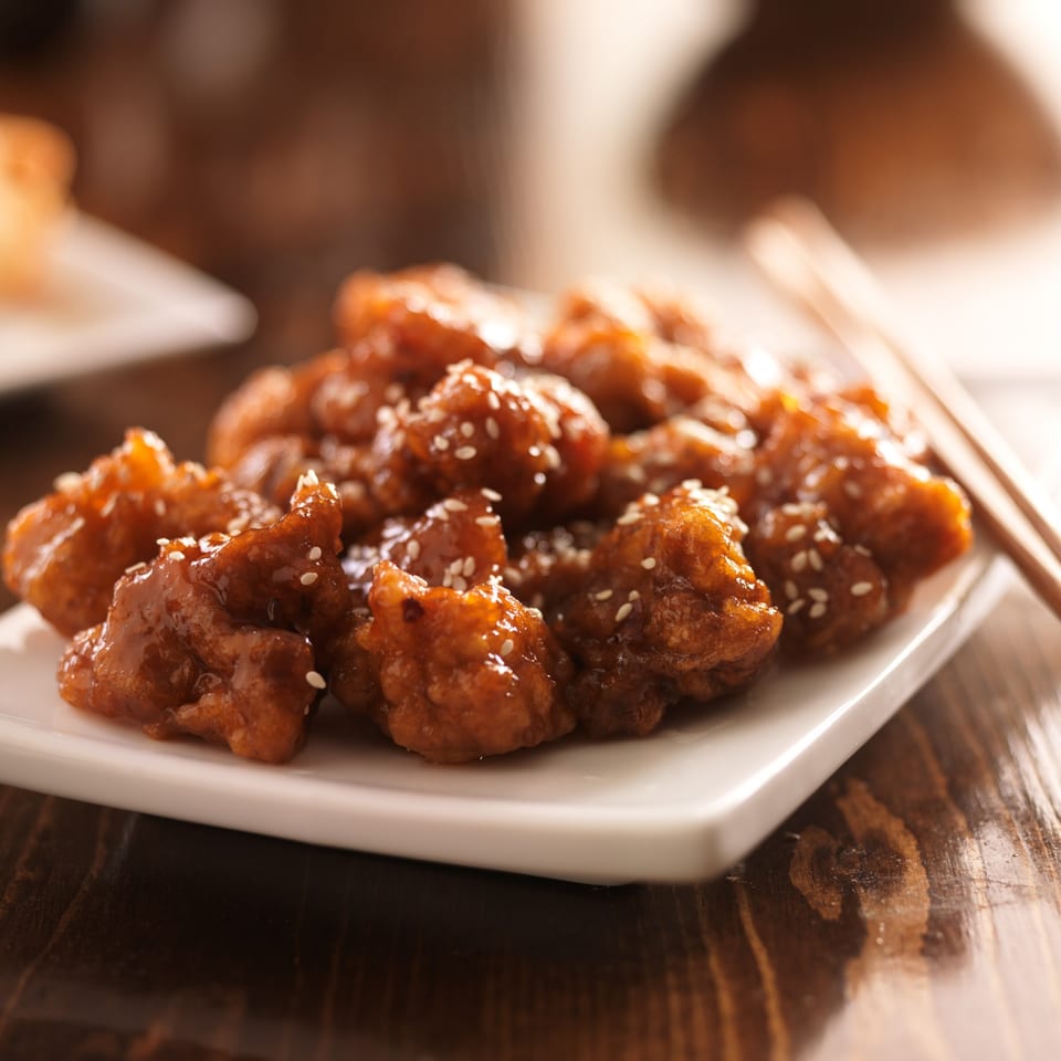 Close-up of sesame chicken pieces on a white rectangular plate with chopsticks.